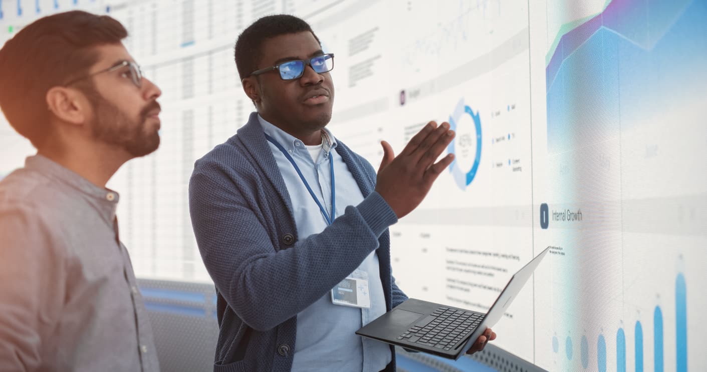 Two men analyze a screen displaying various graphs and data visualizations.